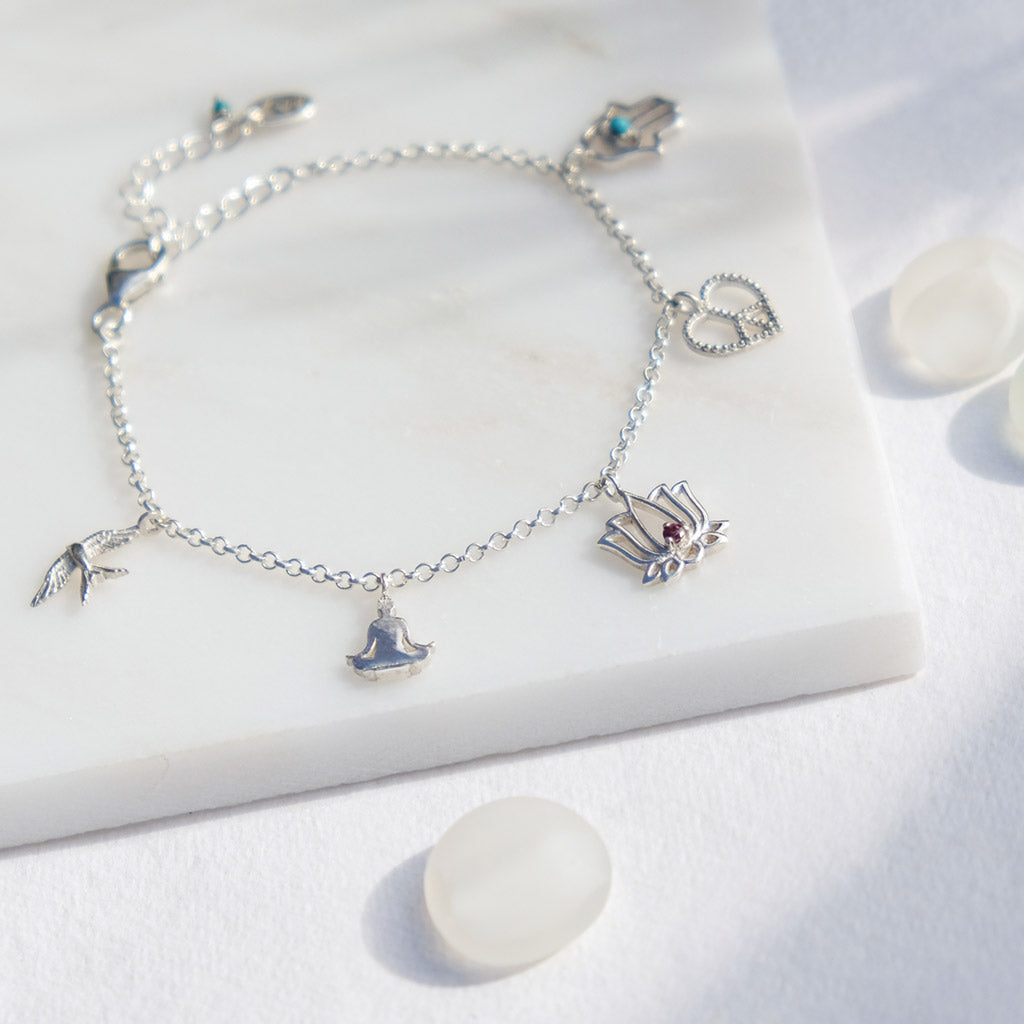 Sterling silver charm bracelet featuring sitting buddha charm, a lotus flower charm with rhodolite garnet, a swallow bird charm to represent freedom, a Hamsa hand charm with turquoise gemstone, and a heart-shaped peace charm against a marble background